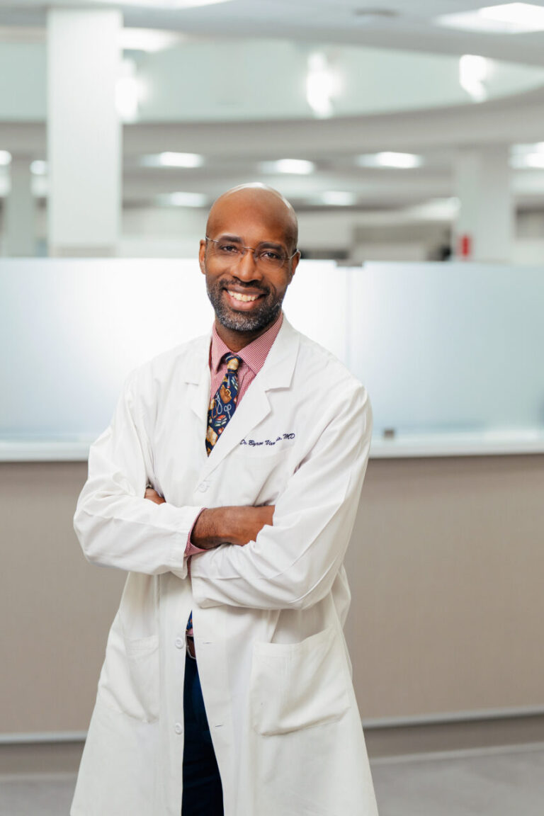 Smiling male doctor in a white coat with arms crossed in a bright medical office.