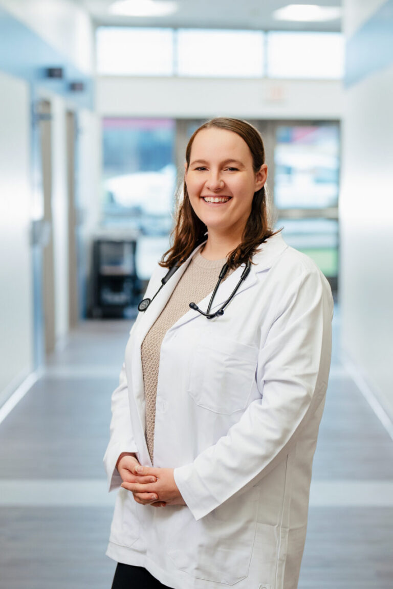 Smiling female doctor in a white coat with a stethoscope stands in a bright hospital hallway.