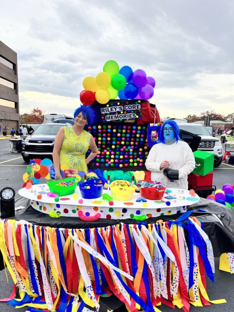 Two people dressed as characters from Inside Out stand behind a colorful table with balloons and decorations labeled "Riley's Core Memories.